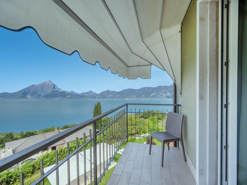 Balcone con sedia, tenda da sole e vista su lago e montagne in giornata limpida.