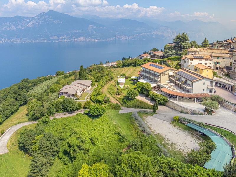 Villaggio di montagna con case e area verde vicino al lago, montagne sullo sfondo sotto un cielo azzurro.