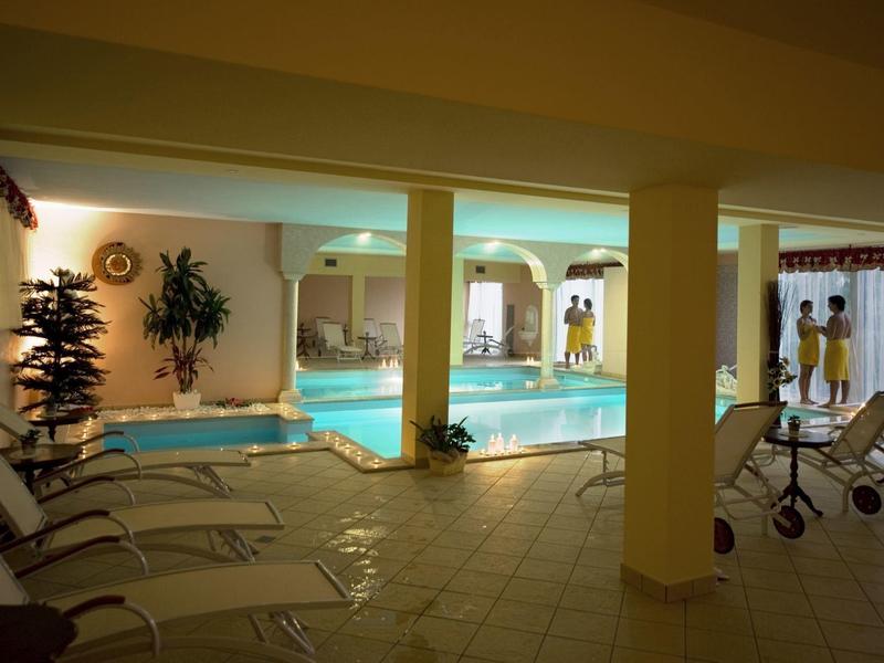 Vue intérieure d'une piscine intérieure avec chaises longues, plantes et personnes en arrière-plan.