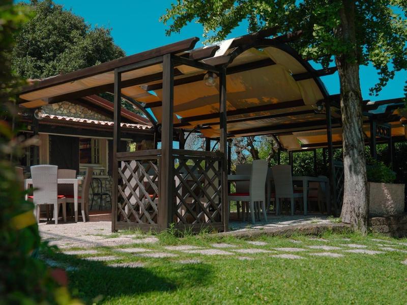 Open paviljoen met tafels en stoelen in een schaduwrijke tuin bij zonlicht.