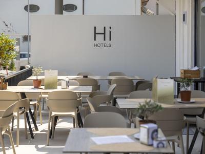 Moderner Außenbereich eines Cafés mit vielen Tischen und Stühlen vor einer Wand mit 'Hi Hotels' Schriftzug.