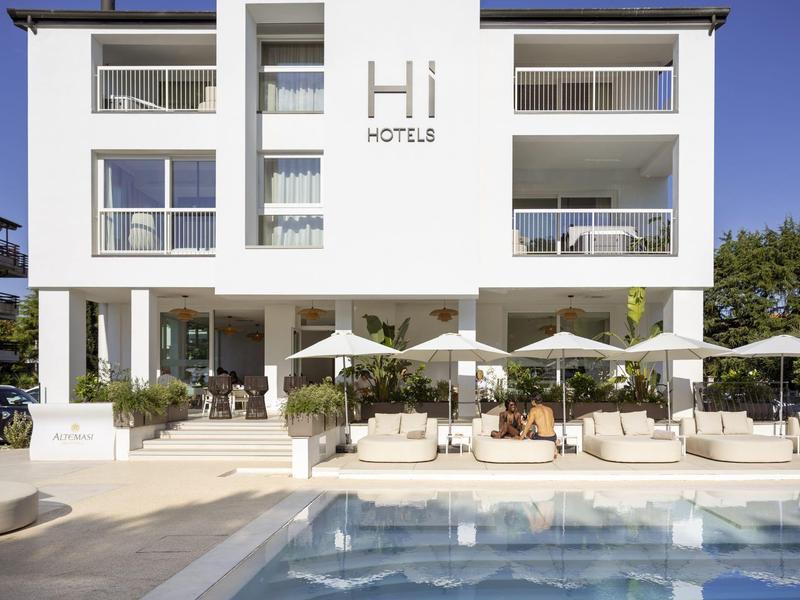 Hotel blanco moderno con piscina, balcones y tumbonas bajo sombrillas en un día despejado.