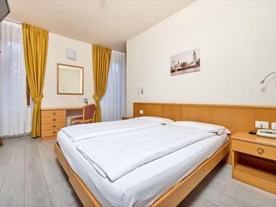 Světly hotelový pokoj s manželskou postelí, dřevěným nábytkem a žlutými závěsy.