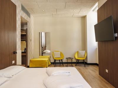 Hotelzimmer mit Doppelbett, zwei gelben Sesseln, Spiegel und Fernseher an der Wand.