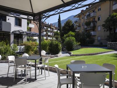 Sitzbereich mit Tischen und Stühlen auf einer Terrasse neben einem gepflegten Garten vor Wohngebäuden.
