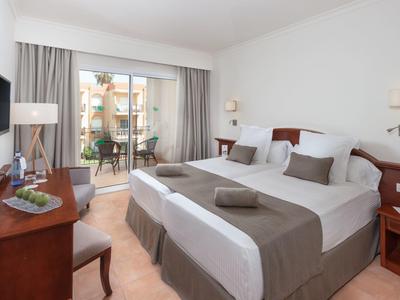 Helles Hotelzimmer mit zwei Einzelbetten, Schreibtisch und Balkon mit Ausblick auf Palmen.