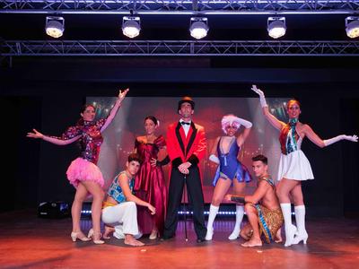 Bunte Gruppe von acht Künstlern in Kostümen auf einer Bühne unter Scheinwerfern.