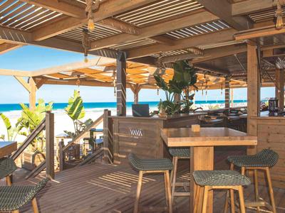 Offenes Strandrestaurant mit Holzmöbeln und Blick auf das Meer bei sonnigem Wetter.