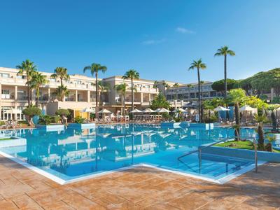 Großer blauer Hotelpool mit Palmen und Liegestühlen unter klarem Himmel.