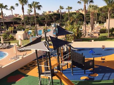 Kinderspielplatz mit Rutschen und Klettergerüsten neben einem Hotelpool und Palmen.