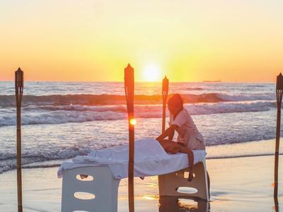 Person, die am Strand bei Sonnenuntergang auf einer Massageliege sitzt, umgeben von Fackeln.