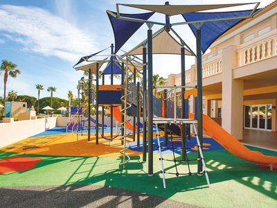 Bunter moderner Spielplatz mit Klettergerüst und Rutschen neben einem Hotelgebäude unter blauem Himmel.