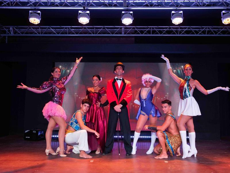 Gruppe von sieben farbenfroh gekleideten Künstlern auf einer Bühne mit schwarzem Hintergrund.