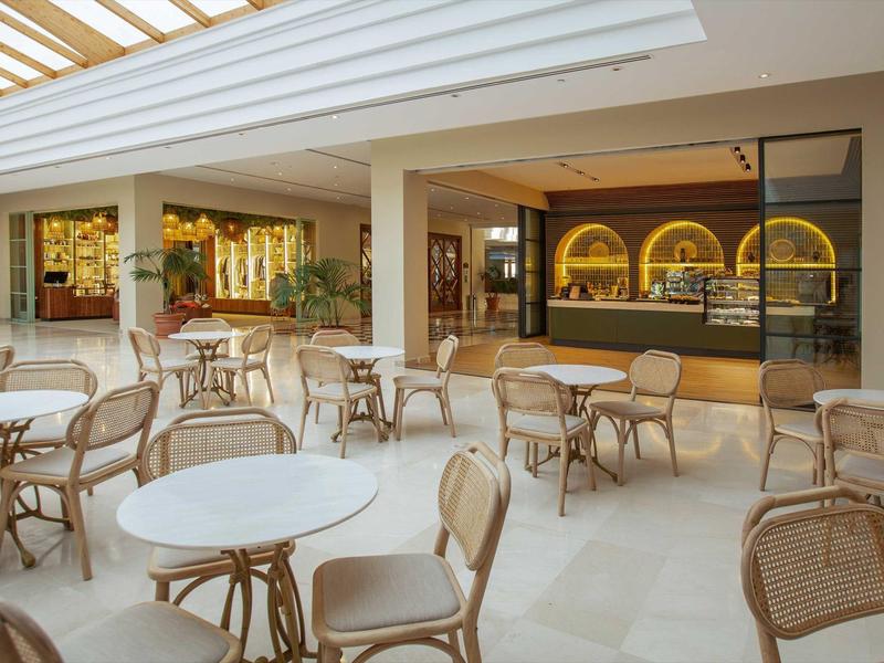 Innenbereich eines Cafés mit weißen Tischen und Stühlen, daneben eine Bar mit gelber Beleuchtung.