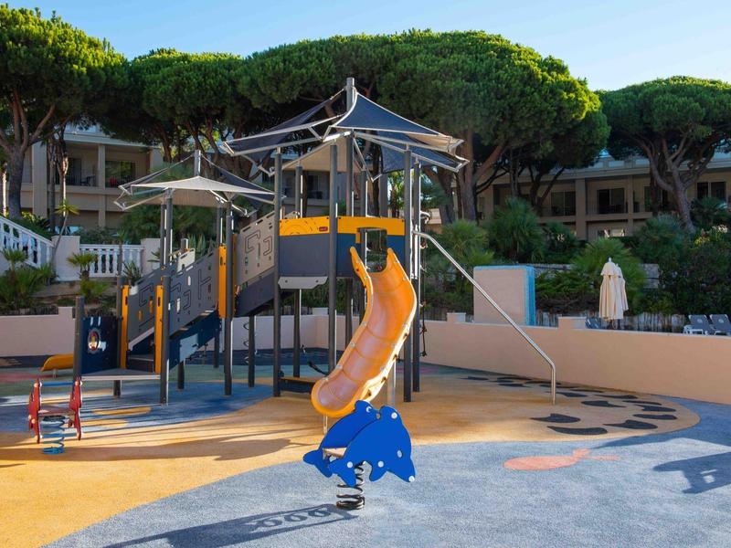 Kinderspielplatz mit Rutsche und Klettergerüst vor Hotelgebäude bei sonnigem Wetter