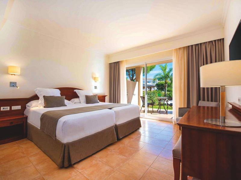 Helles Hotelzimmer mit zwei Betten, Terrasse und Blick auf tropische Pflanzen.