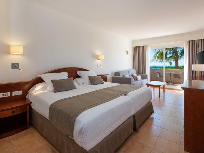 Gemütliches Hotelzimmer mit großen Betten, Sitzbereich und Blick auf Palmen und Meer.