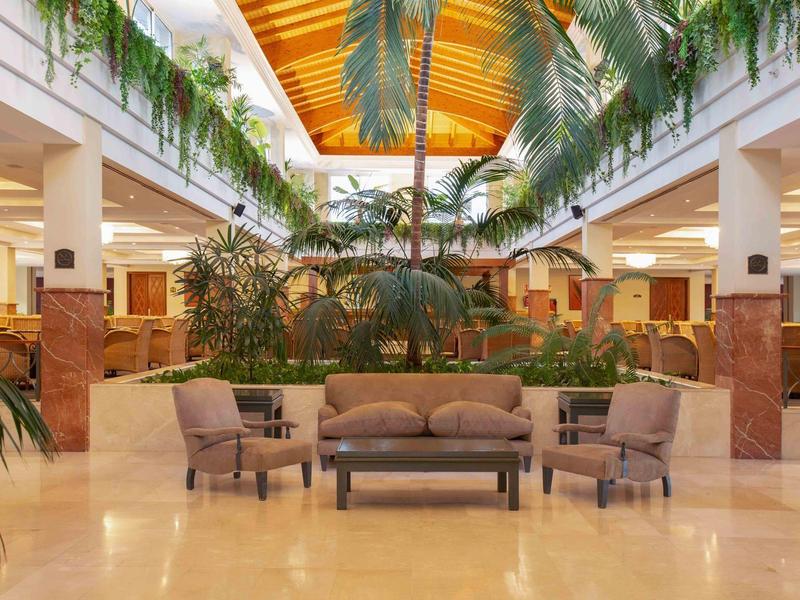 Hotel-Lobby mit gemütlichen Sitzgruppen und grünen Pflanzen unter einem Glasdach.