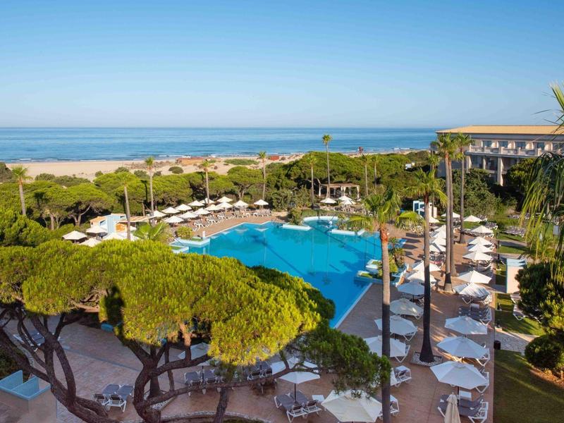 Blick auf einen großen Pool mit weißen Sonnenschirmen und Liegestühlen in einem tropischen Resort am Meer.