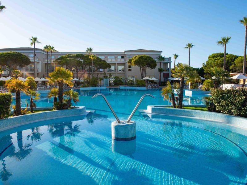 Moderner Außenpool mit Wasserfontänen, umgeben von Palmen und Hotelgebäuden im Hintergrund.