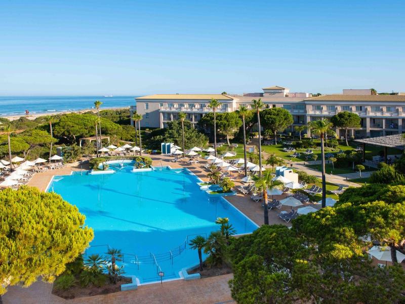 Großer Außenpool mit Liegestühlen und Palmen vor einem Hotel am Meer