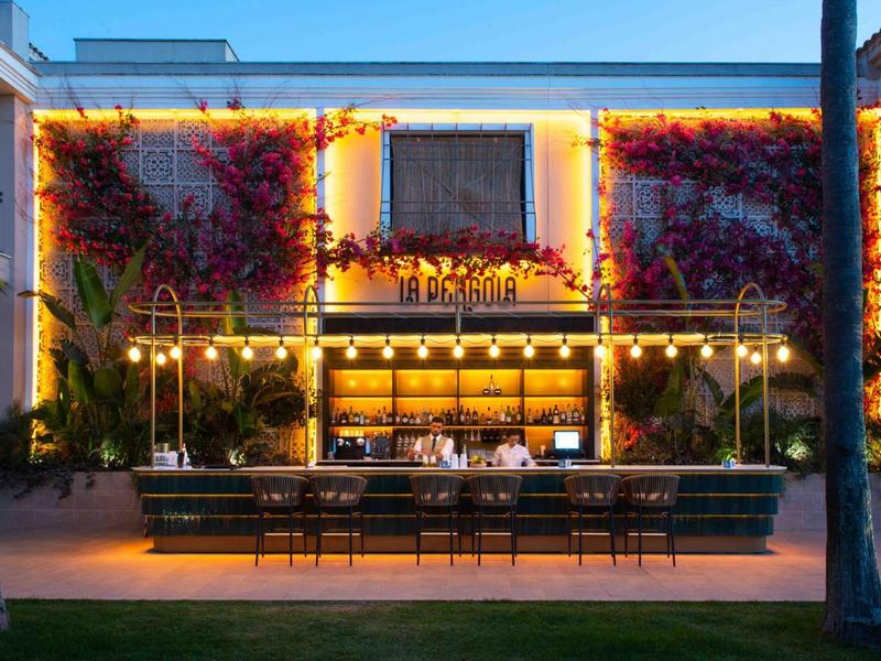 Abendaufnahme einer beleuchteten Open-Air-Bar mit dekorativem Pflanzenbewuchs an der Wand.
