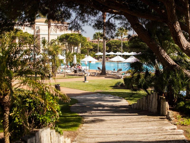 Holzsteg führt durch Schatten zu Poolbereich mit Liegestühlen und Sonnenschirmen in tropischem Garten.
