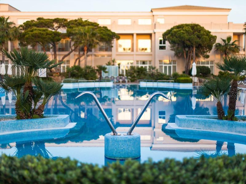 Großer Hotelpool mit Palmen und Liegestühlen vor einem mehrstöckigen Gebäude bei Sonnenuntergang.