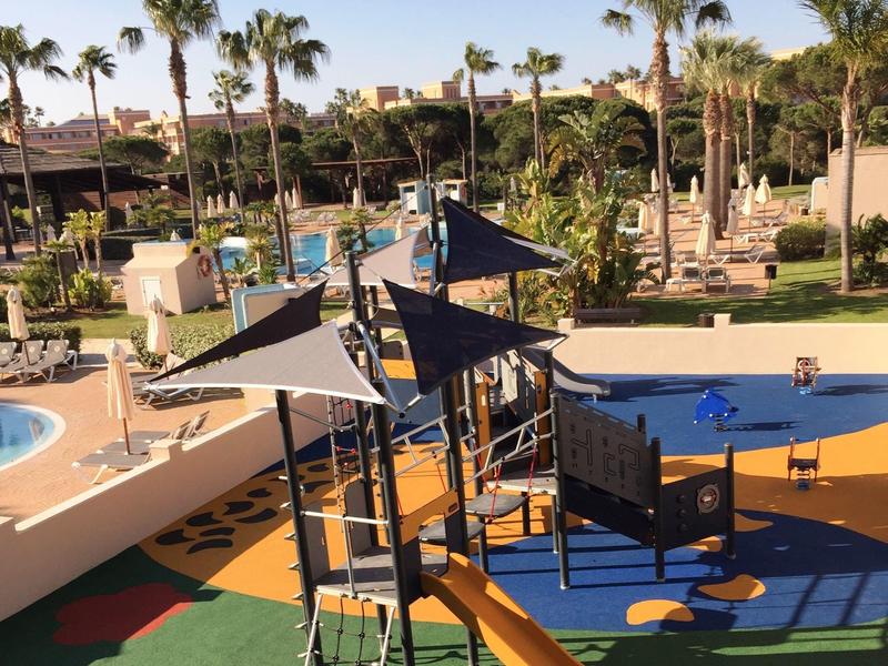 Kinderspielplatz mit Rutschen und Klettergerüsten neben einem Hotelpool und Palmen.