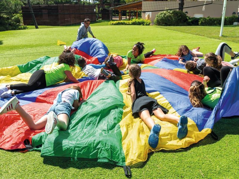 Kinder liegen auf einer bunten Luftmatratze auf einer grünen Wiese neben einem Hotel.