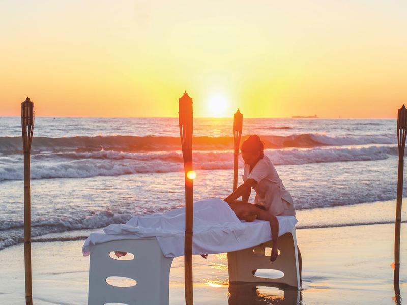 Person, die am Strand bei Sonnenuntergang auf einer Massageliege sitzt, umgeben von Fackeln.