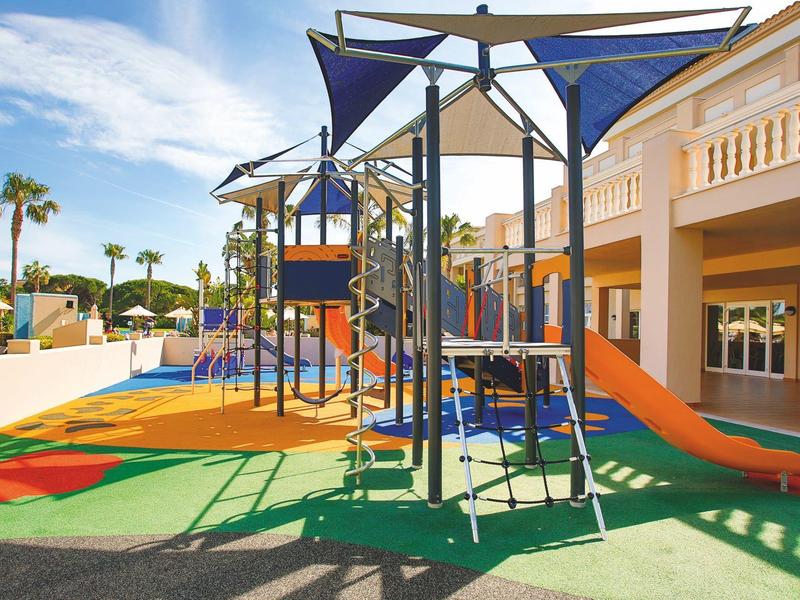 Bunter moderner Spielplatz mit Klettergerüst und Rutschen neben einem Hotelgebäude unter blauem Himmel.