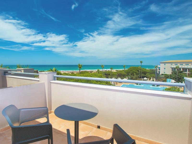 Balkon mit Tisch und Stühlen, Blick auf Pool, Garten und Meer unter blauem Himmel.