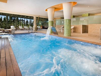 Innen-Schwimmbad mit Holzdeck, Wasserfällen und großen Fenstern mit Blick ins Grüne.