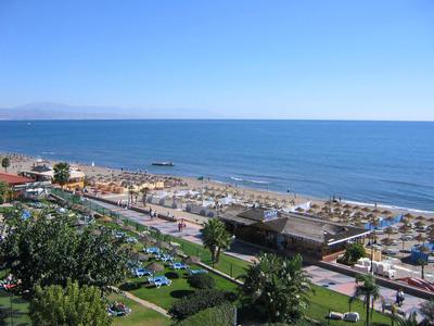 Strand mit Sonnenschirmen, blauem Meer, Promenade und grünen Gärten unter klarem Himmel.