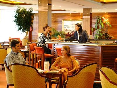 Innenraum eines Cafés mit Gästen an Tischen und einer Kellnerin am Tresen, warme Beleuchtung.