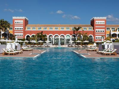 Luxuriöses Hotel mit großem Pool, Liegestühlen und Palmen unter blauem Himmel.