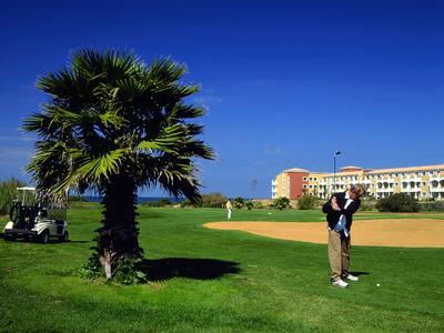 Ein Golfspieler bereitet sich zum Abschlag auf einem grünen Golfplatz vor, Palmen und ein Hotel im Hintergrund.