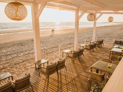 Strandcafé mit Holzboden, Stühlen und Tischen, Lampen hängen von der Decke, Blick auf Meer und Sandstrand.