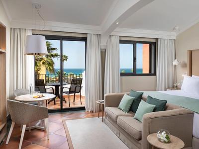 Helles Wohnzimmer mit Sofa, Stuhl, Teppich, großen Fenstern und Balkon mit Meerblick.