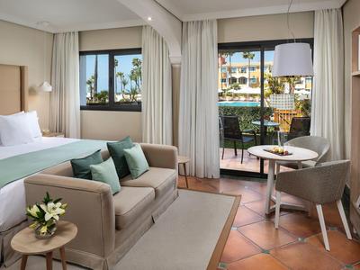 Helles Hotelzimmer mit Bett, Sofa, Sesseln, Terrasse und Blick auf Pool und Palmen.