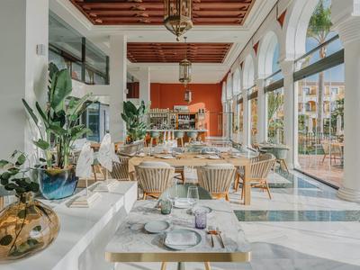 Helles Restaurant mit großen Fenstern, Holztischen, Rattan-Stühlen und grünen Pflanzen.