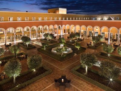 Innenhof mit Brunnen, symmetrischen Beeten, Bäumen und Rosengärten, umgeben von Arkaden bei Abendlicht.