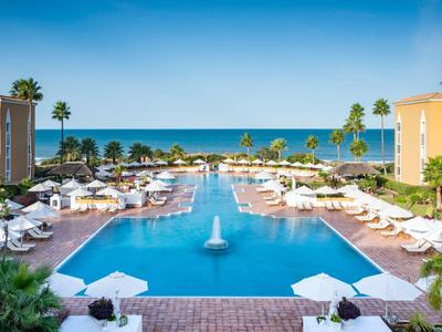 Großer Pool mit Wasserfontäne, umgeben von Liegestühlen und Palmen am Meer unter blauem Himmel.
