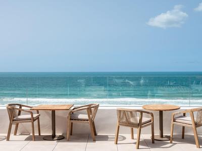 Leerer Balkon mit Holztischen und Stühlen, Blick auf ruhiges türkises Meer und klaren Himmel.