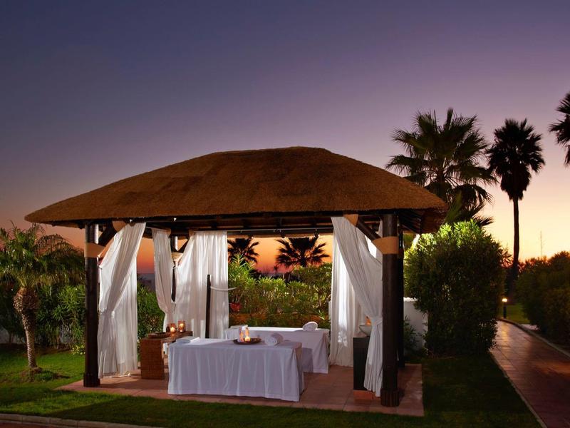 Offener Pavillon mit weißem Vorhang und gedecktem Tisch bei Sonnenuntergang, umgeben von Palmen.