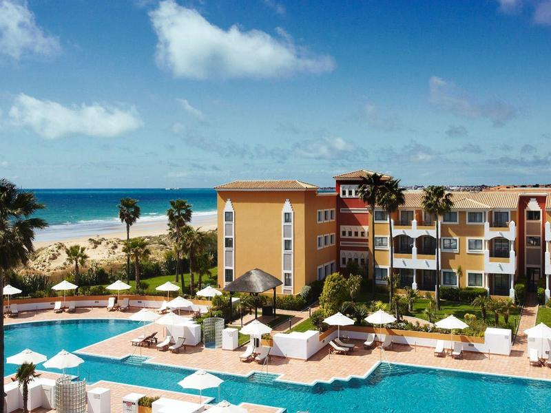 Großes Hotel mit Außenpool, weißen Sonnenschirmen und Blick auf das blaue Meer unter blauem Himmel.