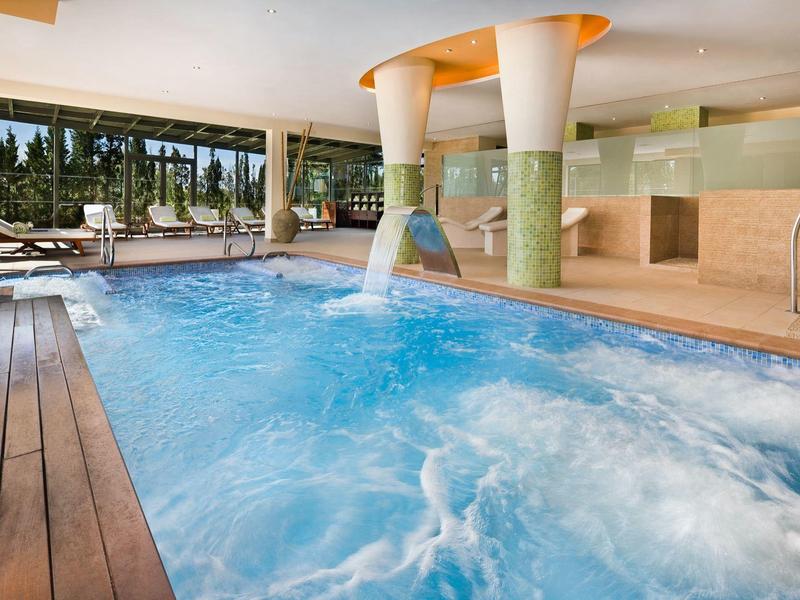 Innen-Schwimmbad mit Holzdeck, Wasserfällen und großen Fenstern mit Blick ins Grüne.