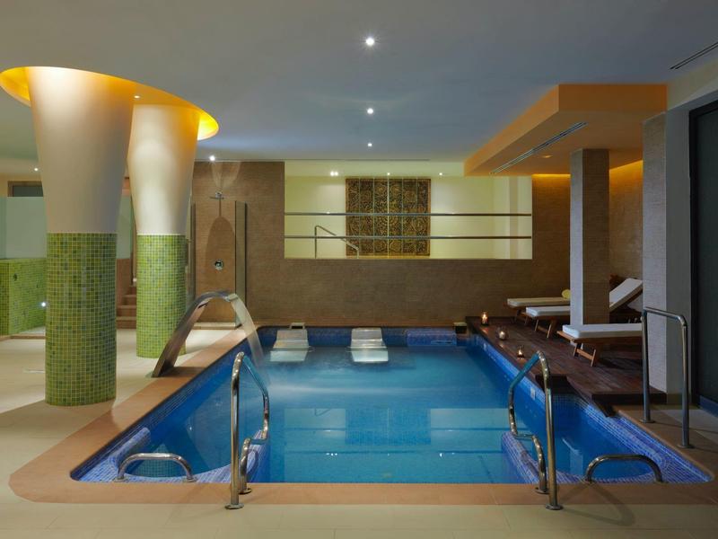 Innenpool mit blauer Wasserfläche, breitem Beckenrand, Liegestühlen und moderner Beleuchtung.