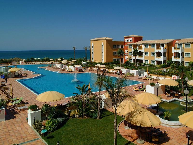 Großer Hotelpool mit Sonnenschirmen, Palmen und Blick aufs blaue Meer unter klarem Himmel.
